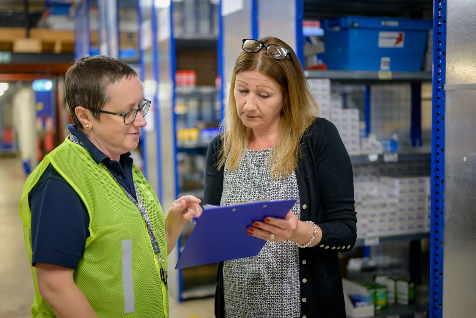 Two women stand in a warehouse aisle between blue shelving; one in a high-visibility vest looks at the notes on the clipboard the other is holding.